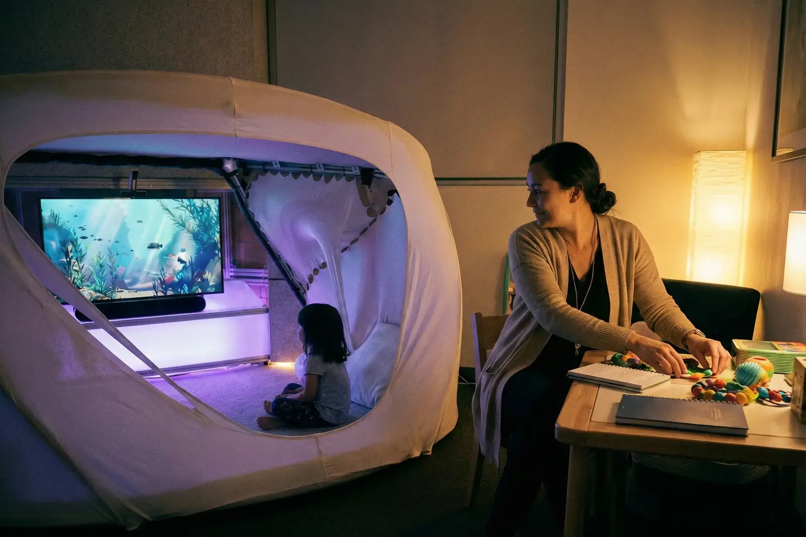 A child in a soft sensory cocoon watching an underwater scene, a parent working at a desk beside it