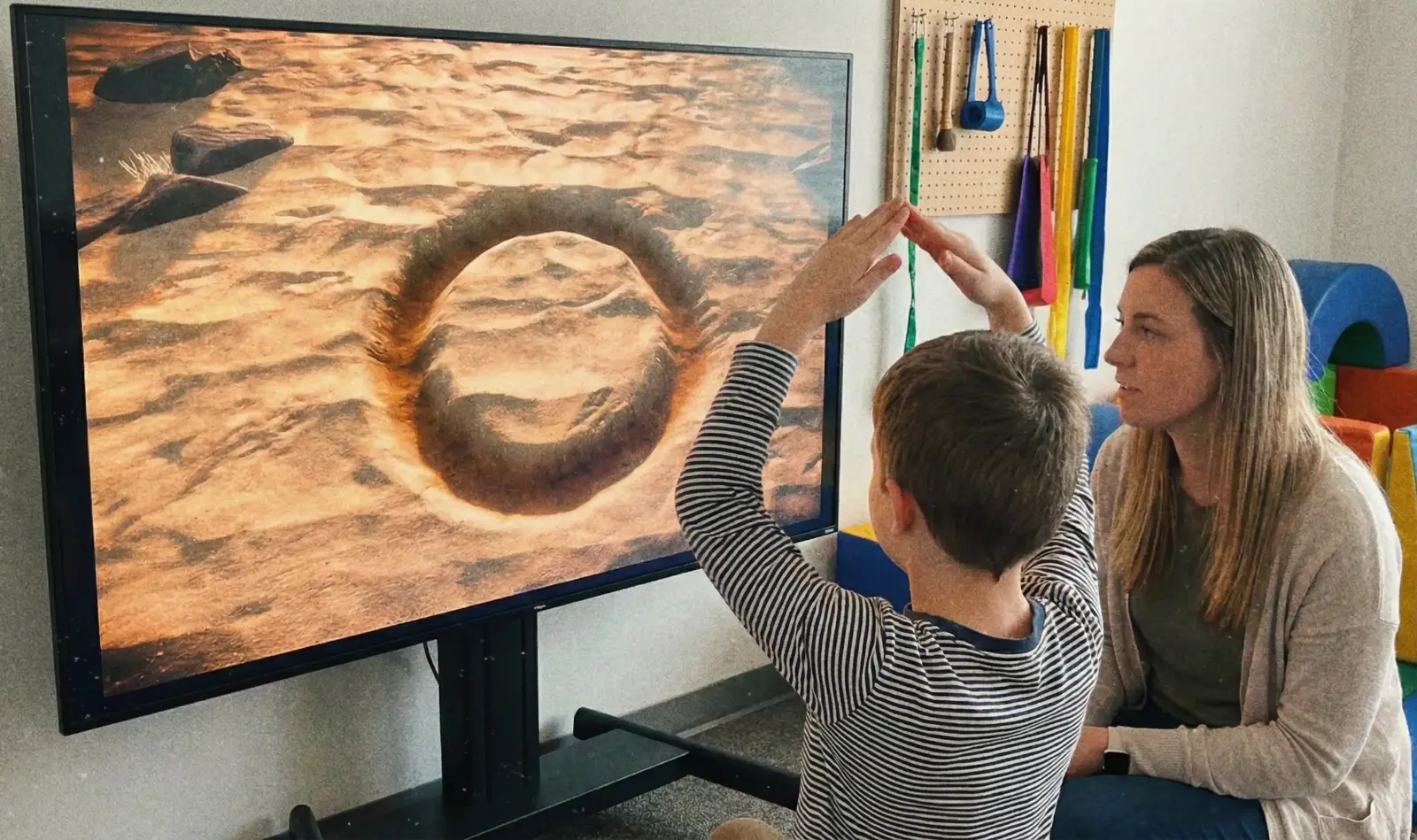 A child traces a circle in the air in front of a large screen, a therapist beside them